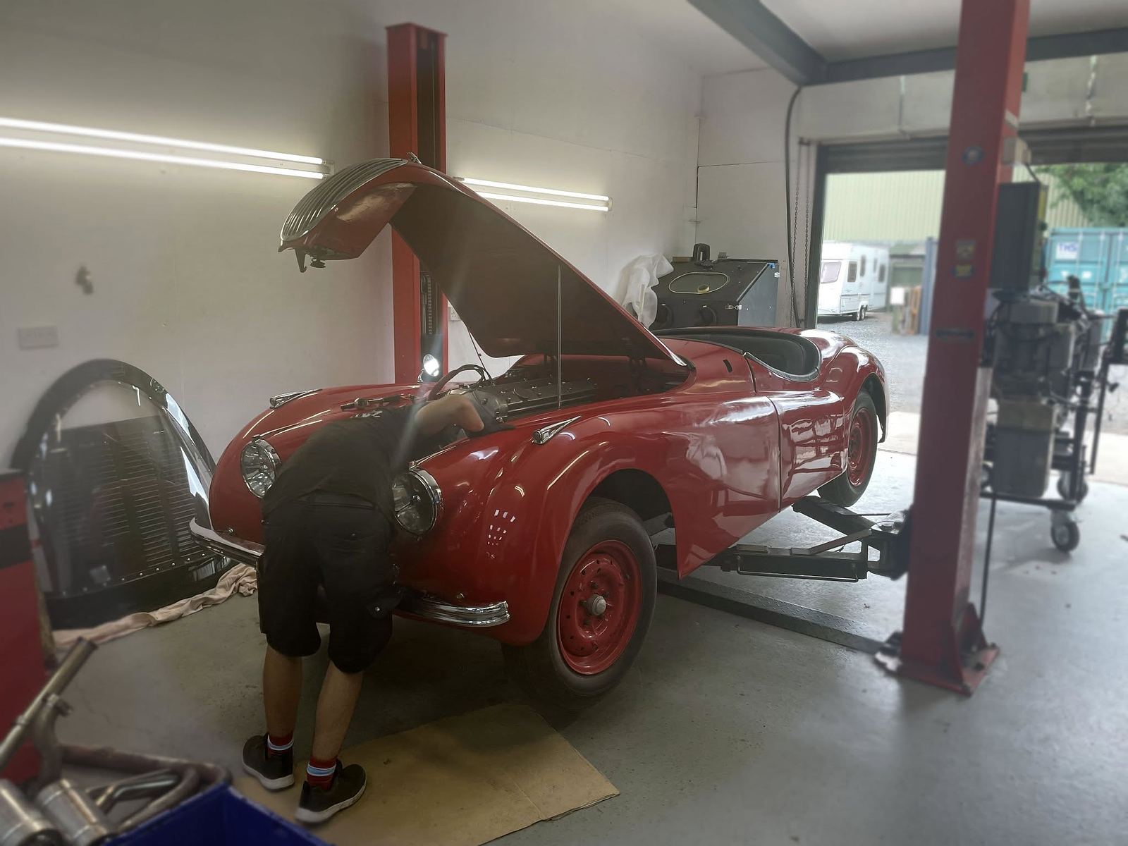 A red classic sports car up on a two-post lift with a mechanic in the engine bay